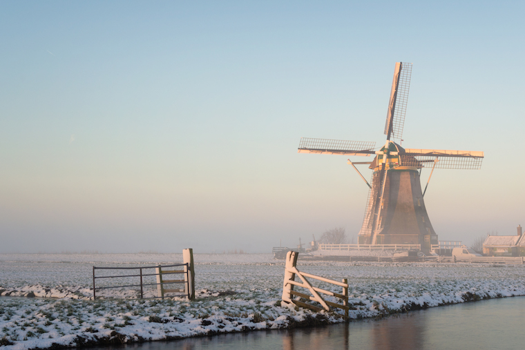 Winter landscape in the Netherlands with a windmill in a meadow, a fence, a canal, snow and fog at sunrise