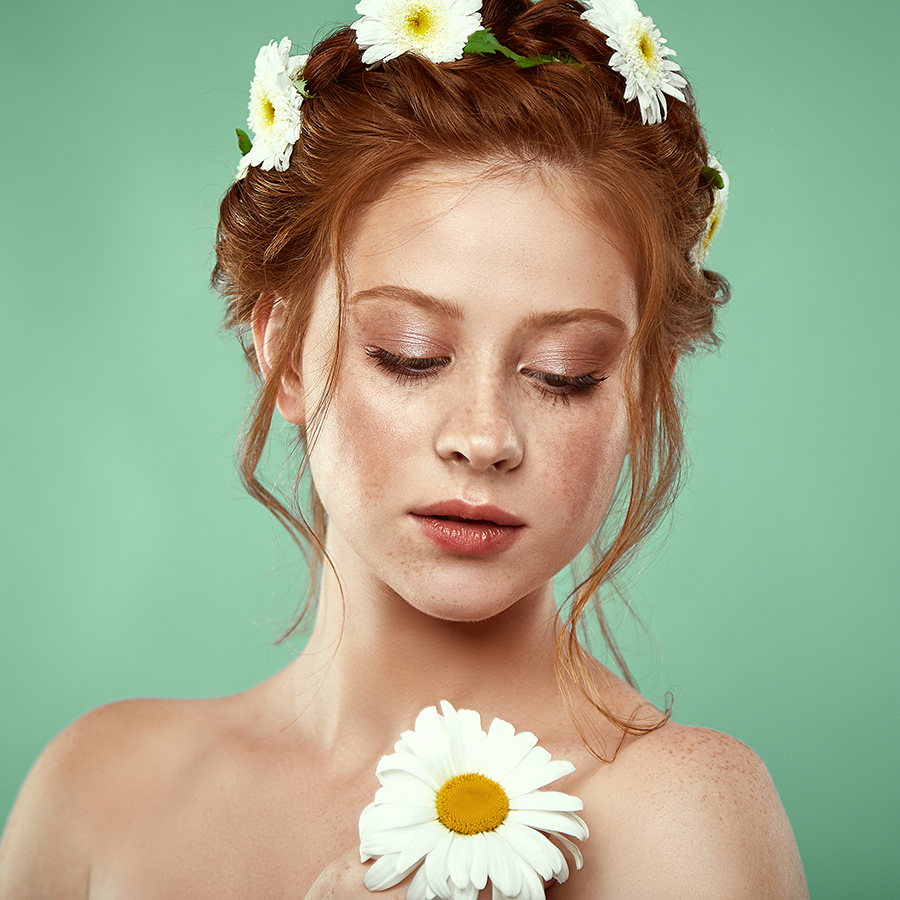 Portrait of beautiful positive redheaded girl with a chamomile crown on her head posing on a bright green studio background