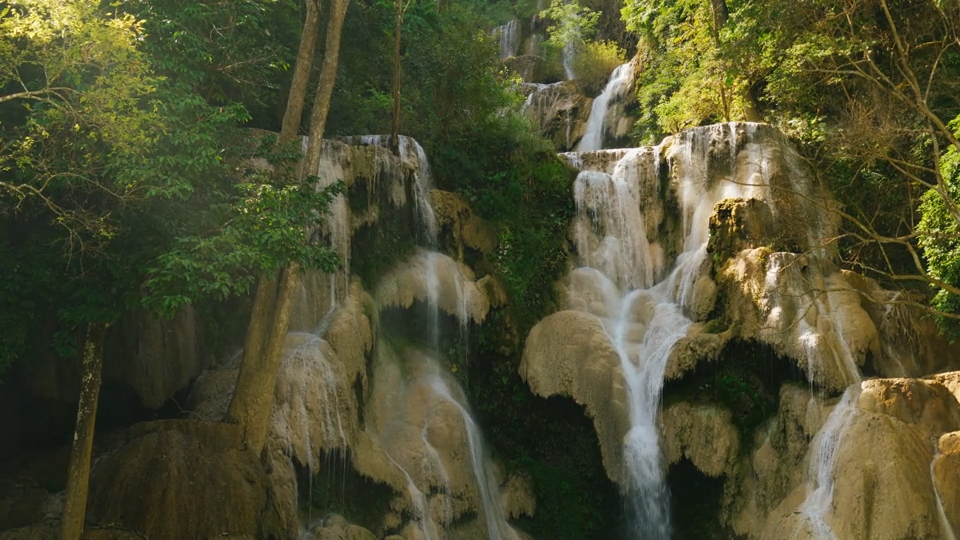 A multi-tiered waterfall with white water flowing over light brown rocks, surrounded by dense green trees and foliage.