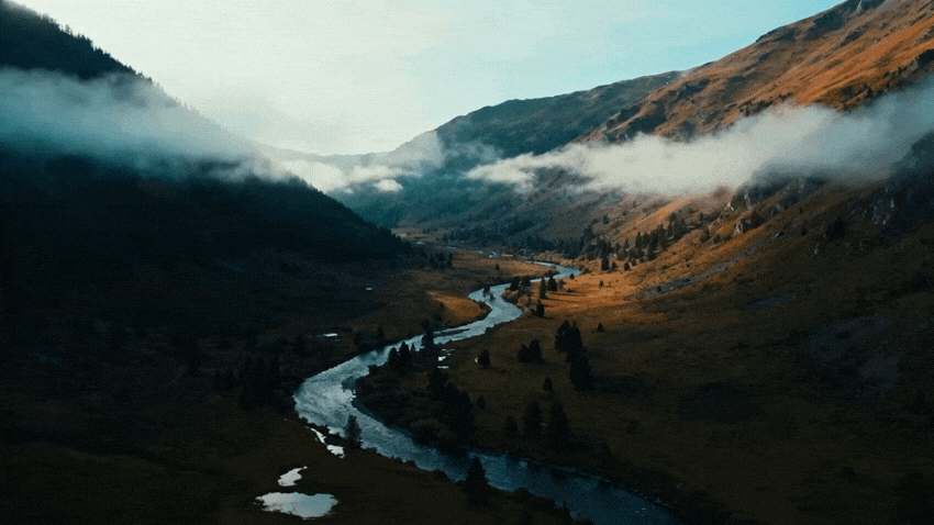 Aerial view of a winding river flowing through a misty mountain valley. One mountain slope is dark and shadowed, while the other is brightly lit by the sun, revealing warm earthy tones.