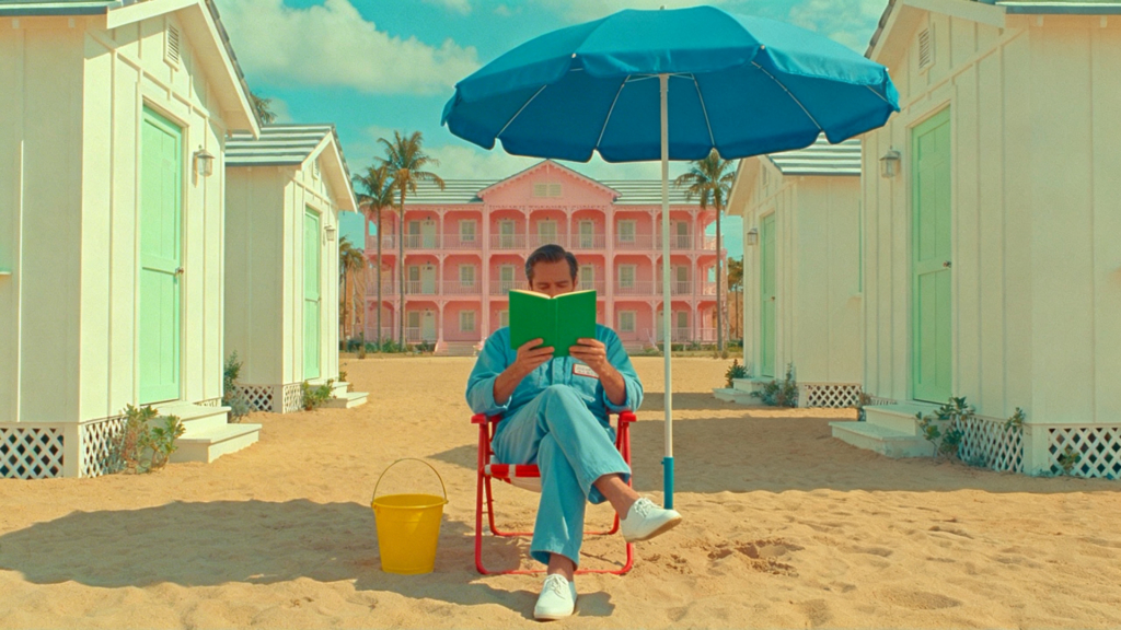 A man in a blue jumpsuit reads a green book under a blue umbrella on a sandy beach. He sits in a red chair. White beach huts with mint green doors flank him, and a large pink hotel stands in the background under a blue sky.