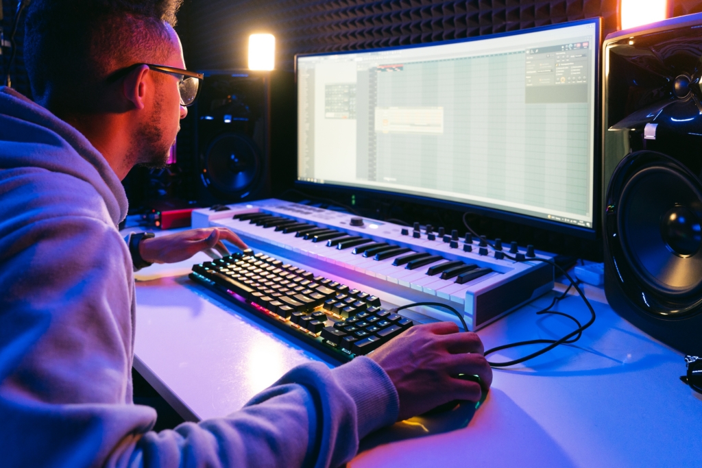 A person in glasses and a hoodie works intently on a computer with a large monitor displaying a music production software (DAW). A MIDI keyboard, an RGB mechanical keyboard, and studio monitors are visible on the desk, all bathed in atmospheric blue and purple lighting.