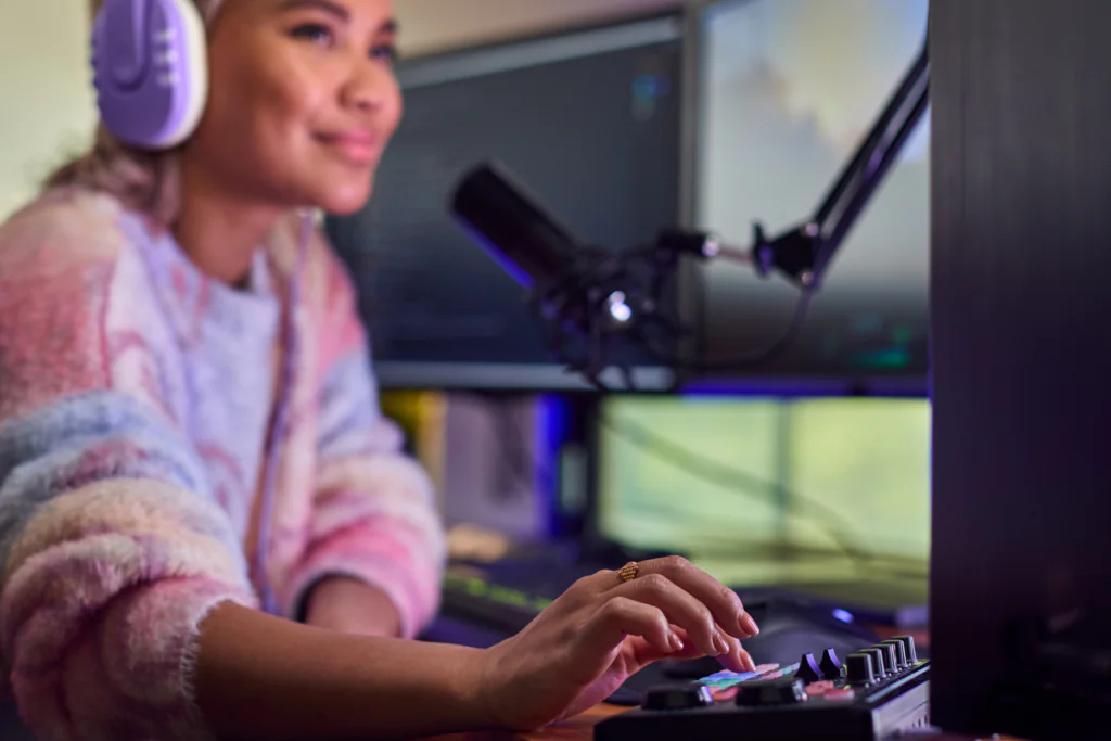 A young woman with purple and white headphones smiles softly while her right hand rests on a control panel with colorful keys. A microphone and computer monitors are visible in the blurred background.