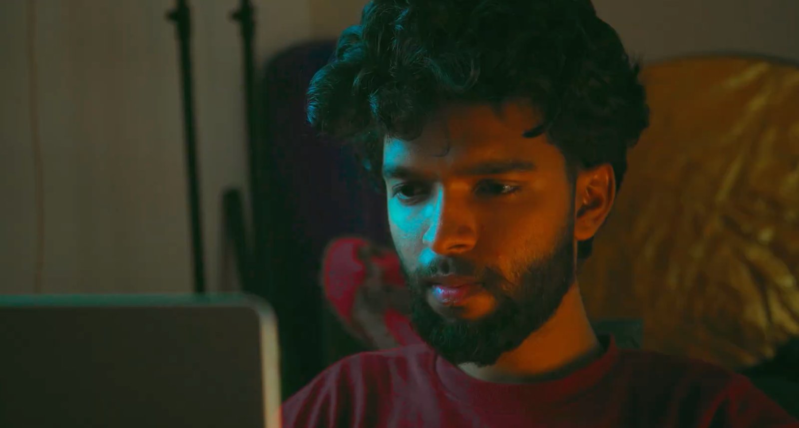 A young man with dark hair and a beard looks intently at a screen, illuminated by blue light from the left and orange light from the right.