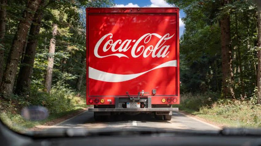 Rear view of a red Coca-Cola truck with its white logo, driving on a paved road surrounded by green trees, seen from a vehicle behind it.