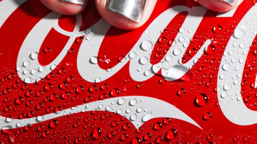 Close-up of a red Coca-Cola can with white logo, covered in water droplets, held by a hand with silver nail polish.