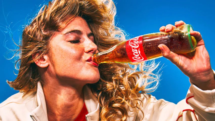 A person with blonde hair and closed eyes drinks from a cold glass Coca-Cola bottle against a blue background.