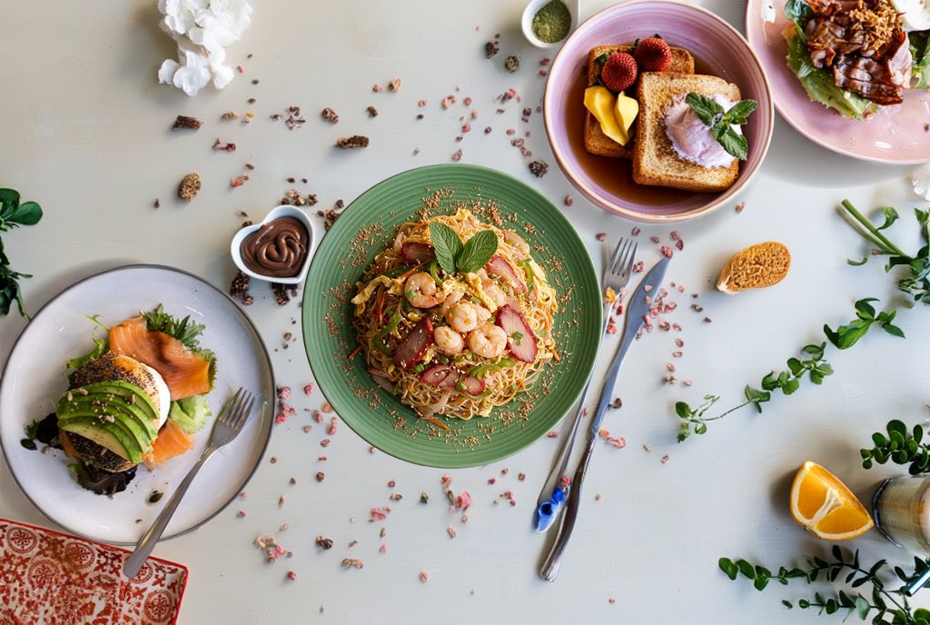 Overhead view of a table laden with various colorful dishes, including shrimp noodles, salmon avocado toast, French toast, and salads.