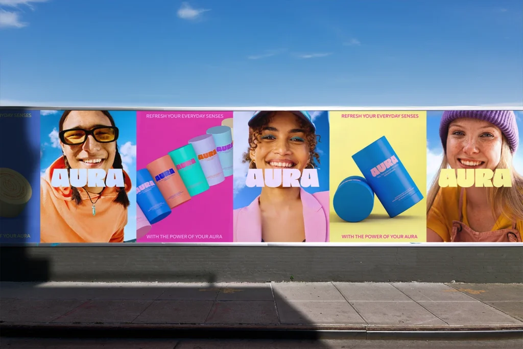 A large outdoor billboard advertising 'AURA' products, featuring five panels. The panels show diverse people smiling, colorful cylindrical products, and text promoting refreshed senses, set against bright backgrounds and a blue sky. The billboard is above a dark brick wall and a concrete sidewalk.