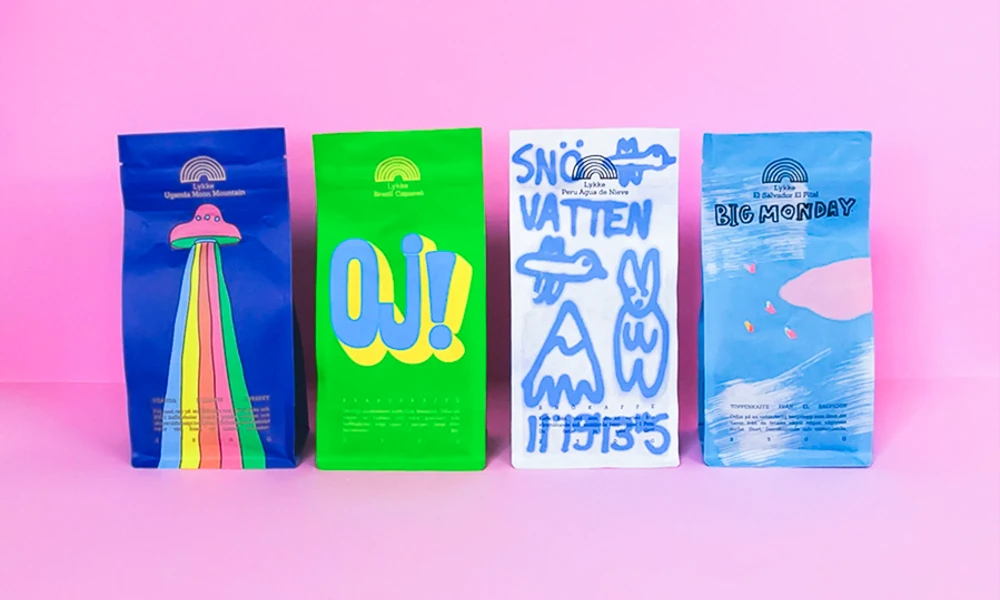 Four colorful Lykke coffee bags standing in a row on a pink surface. Designs include a blue bag with a UFO and rainbow, a green bag with 'OJ!', a white bag with 'SNÖ VATTEN' and doodles, and a light blue bag with 'BIG MONDAY' and a pink cloud.