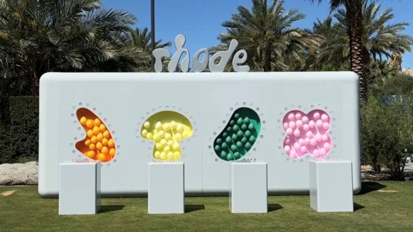A white 'rhode' display with four colorful, sphere-filled cutouts in orange, yellow, green, and pink, on a grassy lawn with palm trees.